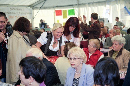 Eine Gruppe von Menschen ist in einem Zelt versammelt, einige stehen und andere sitzen. Eine Frau in traditioneller deutscher Kleidung lächelt und steht neben einer anderen Frau. An der Wand hängen bunte Poster.