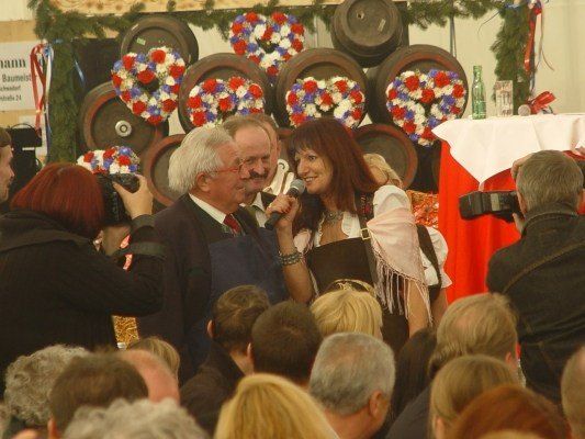 Eine Frau spricht in ein Mikrofon, während sie neben einem Mann in einem Anzug steht, umgeben von einer Menge Menschen. Hinter ihnen befinden sich große Fässer und Kränze.