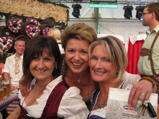 Drei Frauen in traditioneller bayerischer Kleidung lächeln und posieren für ein Foto bei einer Veranstaltung im Freien, wobei ein Mann im Hintergrund einen Schlüsselbund hält.