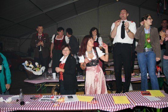 Eine Gruppe von Menschen in traditioneller deutscher Kleidung steht in einem Zelt, einige klatschen und andere stehen hinter Tischen mit rot-weiß karierten Tischdecken. Ein Korb mit Flaschen steht auf einem Tisch.