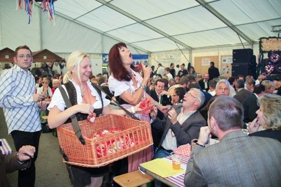 Eine Gruppe von Menschen in traditioneller bayerischer Kleidung versammelt sich in einem Zelt, wobei eine Frau einen Korb mit roten Süßigkeiten hält und eine andere in ein Mikrofon singt.