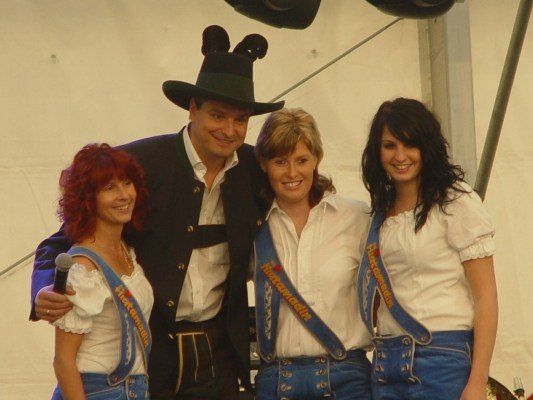 Ein Mann und drei Frauen in traditioneller bayerischer Kleidung stehen zusammen und lächeln für ein Foto. Der Mann trägt einen grünen Hut mit Ohren und hält ein Mikrofon.