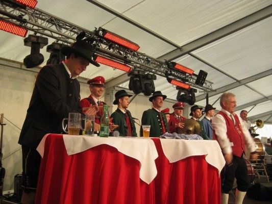 Eine Gruppe von Männern in traditioneller bayerischer Kleidung steht um einen Tisch mit Getränken und Musikinstrumenten. Sie scheinen bei einer Veranstaltung aufzutreten.