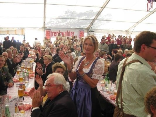 Eine große Menschenmenge in einem Zelt, einige klatschen, eine Frau in traditioneller Kleidung steht vor einem Tisch mit Biergläsern.