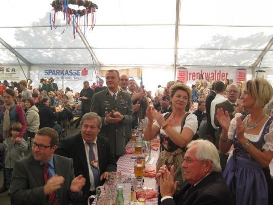 Eine Gruppe von Menschen in formeller Kleidung, einschließlich eines Militäroffiziers, sitzen an einem langen Tisch in einem Zelt, klatschen und lächeln, mit Bierkrügen und Essen auf dem Tisch.