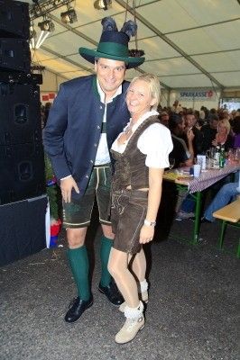 Ein Mann und eine Frau in traditioneller bayerischer Kleidung stehen vor einer Menge auf einem Festival. Der Mann trägt einen grünen Hut und grüne Socken, während die Frau ein braunes Kleid und weiße Schuhe trägt. Hinter ihnen ist ein Tisch mit Flaschen und einer Tischdecke zu sehen.