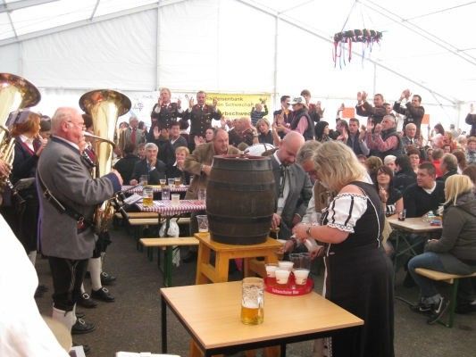Eine große Menschenmenge versammelt sich um einen Tisch mit einem großen Holzfass, eine Frau schenkt Bier in Gläser, während ein Mann Tuba spielt und andere zusehen.