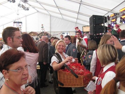 Eine Frau in traditioneller Kleidung hält einen Korb mit roten Süßigkeiten, umgeben von einer Menge bei einem Festival mit Musikern im Hintergrund.