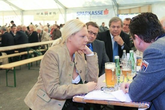 Eine Gruppe von Menschen sitzt an einem Tisch in einem Zelt, wahrscheinlich bei einem Bierfest. Eine Frau schaut einen Mann an, der mit ihr spricht. Auf dem Tisch stehen Gläser mit Bier und Flaschen. Dahinter sitzen weitere Menschen auf Bänken. Ein Banner mit dem Text 'SPARKASSE' ist sichtbar.