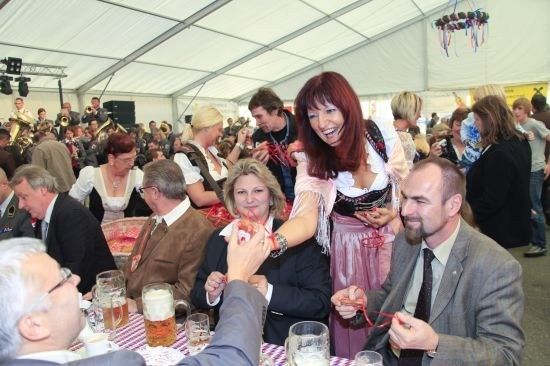 Eine Gruppe von Menschen versammelt sich in einem Zelt, einige tragen traditionelle Kleidung. Eine Frau in einem Dirndl lächelt und hält ein rotes Band, möglicherweise an einer festlichen Veranstaltung teilnehmend.