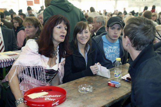 Eine Gruppe von Menschen sitzt an einem Tisch, darunter eine Frau in traditioneller Kleidung, ein Mann mit Kappe und ein Mann in einem blauen T-Shirt. Sie sind von anderen Menschen in einem überfüllten Raum umgeben.