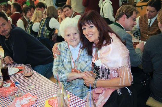 Eine ältere Frau mit weißen Haaren lächelt und hält Händchen mit einer jüngeren Frau in einem rosa Kleid bei einem festlichen Treffen. Sie sind von anderen Menschen umgeben, die an einem Tisch mit Getränken und Essen sitzen.