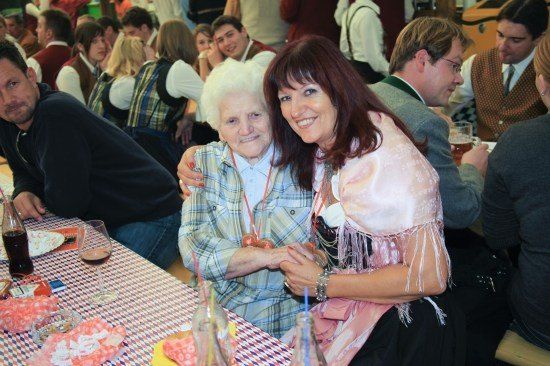 Eine ältere Frau in einem karierten Hemd lächelt und posiert für ein Foto mit einer jüngeren Frau in einem rosa Kleid bei einem festlichen Treffen. Hinter ihnen sitzen mehrere Personen an einem Tisch, einige halten Gläser mit Bier.