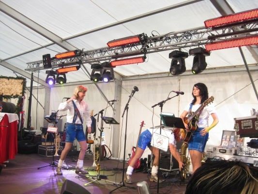Eine Band tritt auf einer Bühne unter einem Zelt auf, mit zwei Gitarristen, einem Trompeter und einem sitzenden Saxophonisten. Die Bühne ist mit roten Lichtern beleuchtet und hat verschiedene Musikinstrumente und Ausrüstung im Hintergrund.