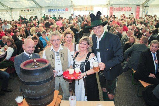 Eine Gruppe von Menschen in traditioneller deutscher Kleidung posiert für ein Foto mit einem großen Bierfass und Bierkrügen in einem überfüllten Bierzelt.