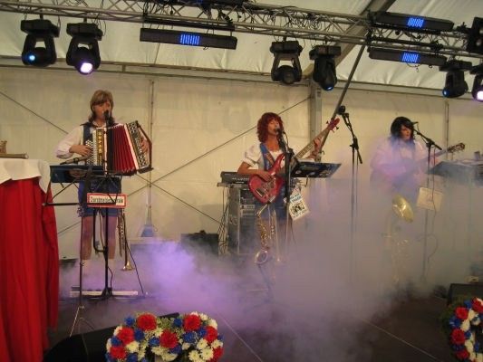 Drei Frauen spielen auf der Bühne mit Musikinstrumenten, darunter Akkordeon, Gitarre und Trompete, unter Scheinwerfern mit Raucheffekten.