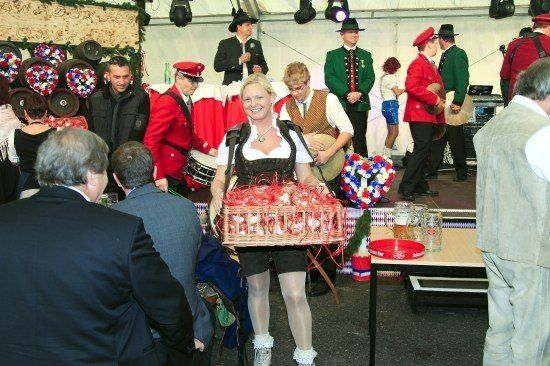 Eine Frau in traditioneller bayerischer Kleidung hält einen Korb mit roten Gegenständen, während sie vor einer Gruppe von Menschen in roten und grünen Uniformen steht. Sie befinden sich auf einer Bühne mit Musikinstrumenten und Dekorationen.