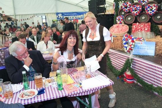 Eine Gruppe von Menschen versammelt sich bei einer Outdoor-Veranstaltung. Zwei Frauen lächeln und stehen neben einem Tisch mit Getränken und Essen. Ein Mann sitzt am Tisch.