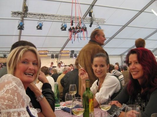 Drei Frauen sitzen an einem Tisch in einem Zelt, lächeln und trinken Wein. Ein Mann geht hinter ihnen, und es gibt andere Menschen, die sitzen und stehen. Das Zelt hat eine weiße Decke und Metallträger mit Lichtern.