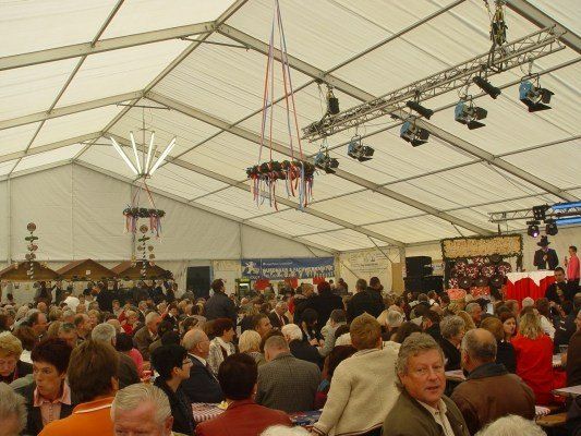 Eine große Menschenmenge versammelt sich in einem Zelt, wahrscheinlich für ein Festival. Das Zelt ist mit hängenden Lichtern und Bannern dekoriert. Einige Menschen stehen auf der Bühne, andere sitzen an Tischen.