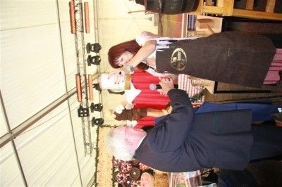 Ein Mann in einem Anzug überreicht einer Frau in einem roten Kleid und einer Schürze eine Auszeichnung, mit einer Statue im Hintergrund.