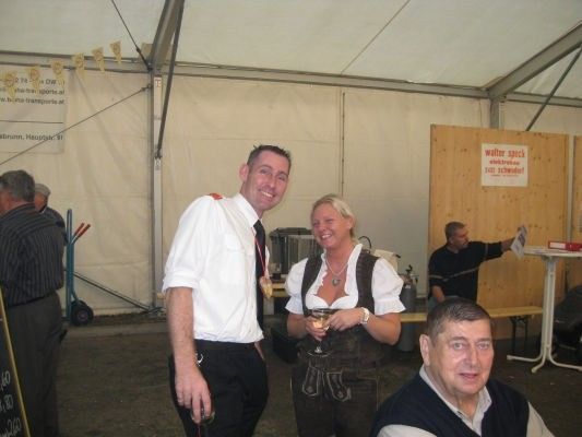 Ein Mann und eine Frau in traditioneller bayerischer Kleidung stehen lächelnd in einem Zelt mit anderen Leuten. Die Frau hält ein Weinglas, und ein Mann sitzt auf einer Bank hinter ihnen. Eine Holztür mit einem Schild steht rechts.