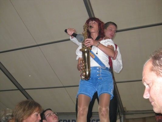 Eine Frau in einem weißen Oberteil und blauen Shorts spielt Saxophon und hält ein Mikrofon, während ein Mann sie von hinten unterstützt.