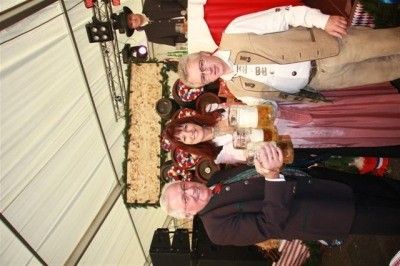 Eine Gruppe von Menschen in traditioneller Kleidung posiert für ein Foto bei einer Veranstaltung und hält große Bierkrüge in den Händen. Der Hintergrund zeigt eine festliche Umgebung mit Dekorationen und Beleuchtung.