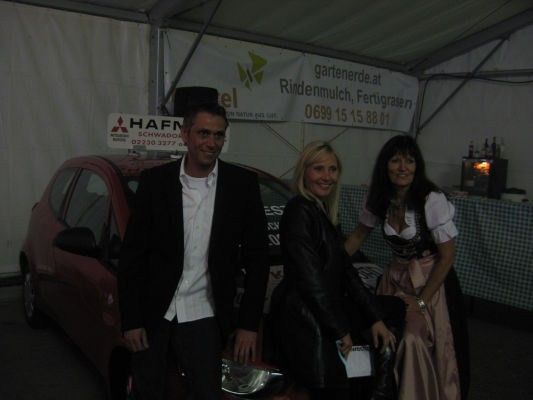 Drei Personen stehen neben einem roten Auto in einem Zelt, wobei eine Frau ein Papier hält und lächelt. Der Mann links trägt einen schwarzen Anzug, und die Frau rechts trägt ein traditionelles deutsches Kleid. Ein Banner mit einer Telefonnummer ist im Hintergrund sichtbar.