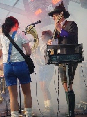 Eine Band spielt auf der Bühne, eine Frau in blauen Shorts und ein Mann in traditioneller bayerischer Kleidung spielt Gitarre und Keyboard.