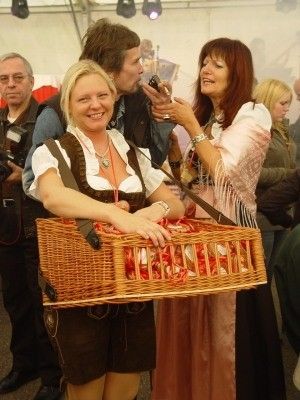 Eine Frau in traditioneller bayerischer Kleidung hält einen Korb voller rot verpackter Süßigkeiten und lächelt, während eine andere Frau in einem rosa Kleid eine Kamera hält. Hinter ihnen versammeln sich Menschen, einige tragen Brillen und Hüte.