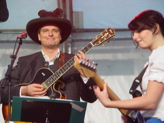 Ein Mann mit Cowboyhut und eine Frau in einem weißen Oberteil spielen Gitarren auf der Bühne. Der Mann lächelt und schaut die Frau an.