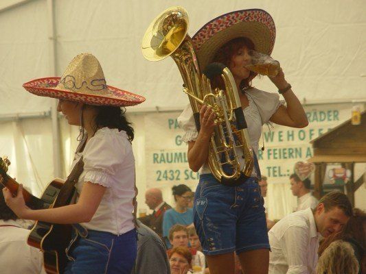 Zwei Frauen in Sombreros, eine spielt Gitarre, die andere spielt Tuba und trinkt ein Bier, in einer festlichen Outdoor-Umgebung.