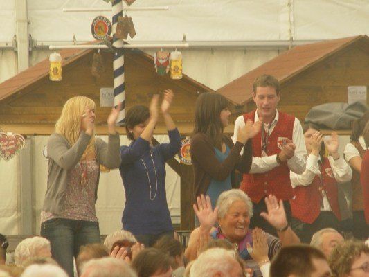 Eine Gruppe von Menschen klatscht und lächelt in einer festlichen Umgebung, möglicherweise bei einem Bierfest. Eine Frau in einer roten Weste steht vor ihnen.