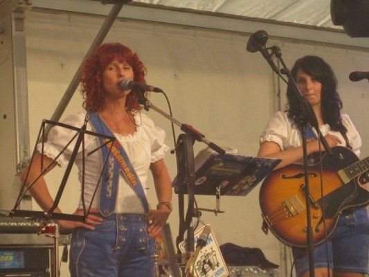 Zwei Frauen treten auf der Bühne auf; eine singt in ein Mikrofon, während die andere Gitarre spielt. Sie tragen passende weiße Blusen und Jeansshorts.