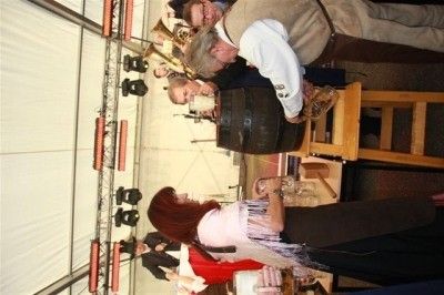 Eine Band spielt auf einer Bühne mit einer Frau in einem roten Kleid und einem Mann in einem weißen Hemd. Die Bühne ist mit Scheinwerfern und einem Holzstuhl ausgestattet.