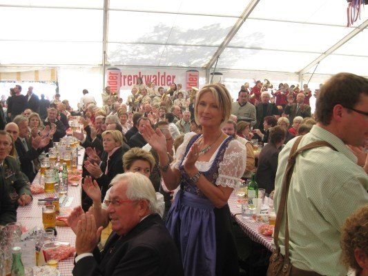 Eine große Gruppe von Menschen ist in einem Zelt zu einer Feier versammelt. Eine Frau in traditioneller deutscher Kleidung klatscht. Viele Menschen sitzen an langen Tischen mit Gläsern Bier.