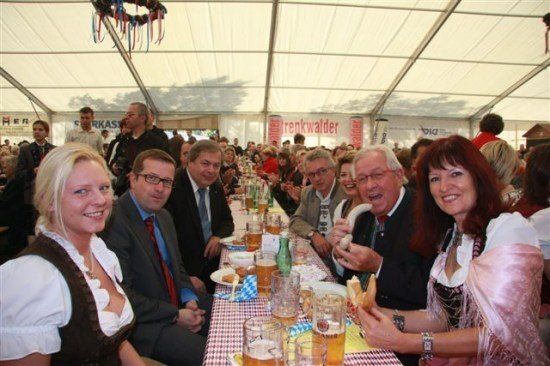 Eine Gruppe von Menschen in traditioneller Kleidung sitzt an einem langen Tisch in einem Zelt, lächelt und posiert für ein Foto. Sie sind von Tellern, Gläsern und Essen umgeben. Das Zelt hat Banner und eine weiße Decke.