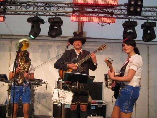 Eine Band spielt auf der Bühne mit einem Mann, der Gitarre spielt, einer Frau, die Bassgitarre spielt, und einem weiteren Mann, der Tuba spielt. Die Bühne ist mit Beleuchtung und einem weißen Hintergrund ausgestattet.