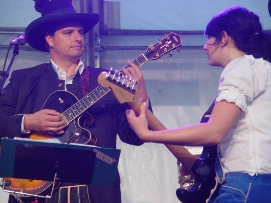 Ein Mann in einem schwarzen Anzug und Hut spielt Gitarre, während eine Frau in einem weißen Oberteil und blauer Jeans ihm auf der Bühne hilft.