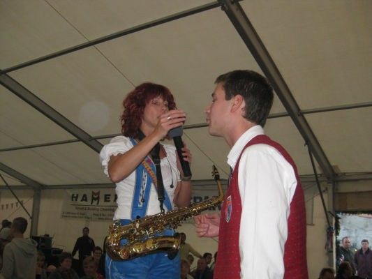 Eine Frau singt in ein Mikrofon, während sie eine Saxophon hält, und ein Mann steht neben ihr, möglicherweise spielt er das Saxophon. Sie treten vor einem Publikum auf.