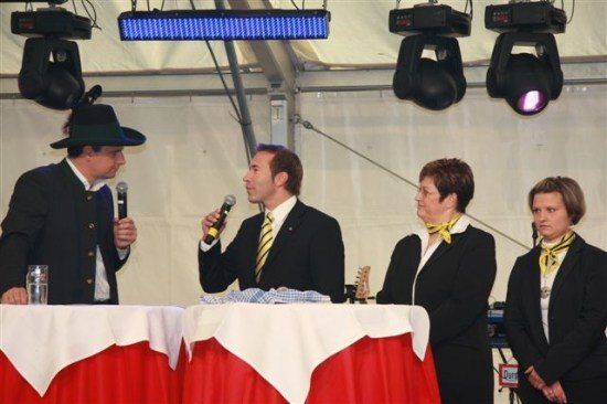 Vier Personen stehen an einem Tisch, zwei Männer und zwei Frauen. Der Mann in der Mitte hält ein Mikrofon und der Mann links trägt einen Hut. Die Frauen tragen gelbe Schals. Scheinwerfer sind an der Decke montiert.