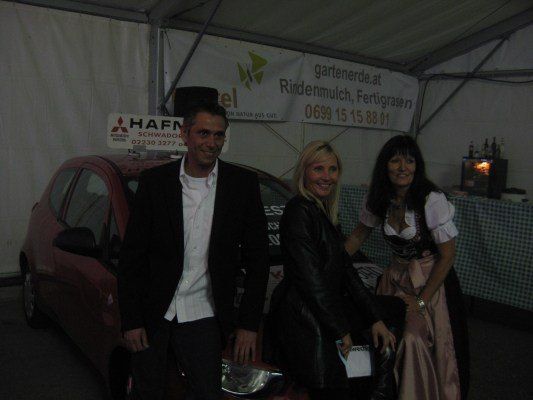 Drei Personen stehen neben einem roten Auto mit einem Schild, auf dem 'Hafen' steht. Der Mann trägt einen Anzug, und die Frauen tragen eine Lederjacke und ein traditionelles Kleid. Sie stehen unter einem Zelt mit einem Banner, das 'gartenerde.at' bewirbt.