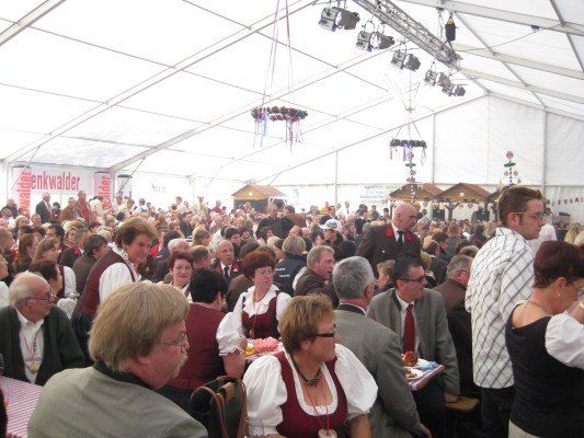 Eine große Gruppe von Menschen ist in einem Zelt versammelt, wahrscheinlich für ein Festival. Einige sitzen, andere stehen und einige tragen traditionelle Kleidung. Das Zelt hat eine weiße Decke und hängende Dekorationen.