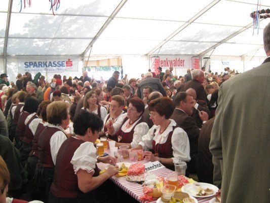 Eine große Gruppe von Menschen in traditioneller deutscher Kleidung sitzt an einem langen Tisch in einem Zelt und genießt Essen und Getränke. Das Zelt hat Banner mit Logos und Text.