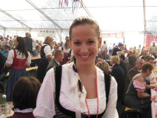 Eine Frau in traditioneller bayerischer Kleidung steht in einem überfüllten Zelt und lächelt in die Kamera. Hinter ihr sitzen eine große Gruppe von Menschen an Tischen und genießen Essen und Getränke.