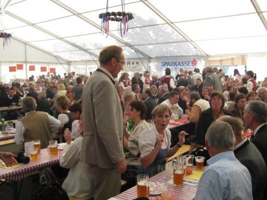Eine große Menschenmenge sitzt in einem Zelt an Tischen, einige in traditioneller Kleidung. Ein Mann in Anzug steht vor einem Tisch mit Biergläsern.