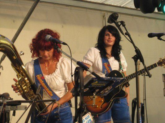 Zwei Frauen spielen auf der Bühne, eine spielt Saxophon und die andere Gitarre. Sie tragen beide weiße Blusen und blaue Jeansshorts.