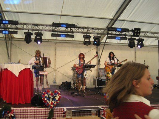 Eine Band spielt auf einer Bühne mit einem rot-weißen Tischtuch, einem Blumenstrauß und einer zuschauenden Frau. Die Band besteht aus drei Mitgliedern, einem spielt Akkordeon, einer spielt Gitarre und einer spielt Saxophon. Die Bühne ist von Scheinwerfern beleuchtet.
