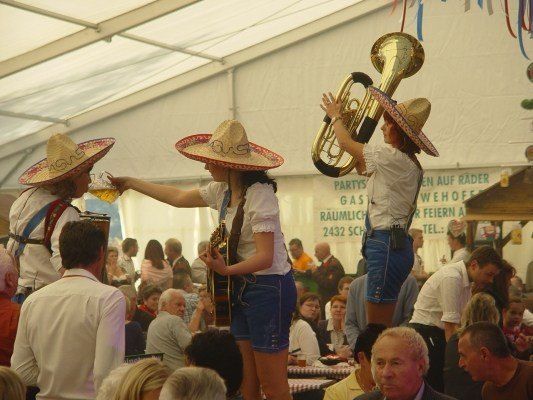 Eine Gruppe von Menschen in Sombreros und Jeansshorts tritt auf der Bühne bei einer Outdoor-Veranstaltung auf, einer spielt Gitarre und der andere hält eine Tuba. Das Publikum sitzt vor ihnen.